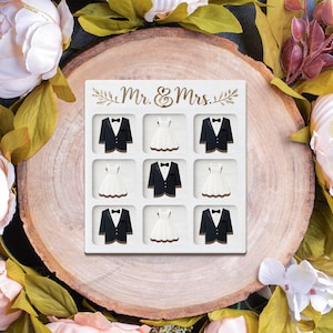 May include: A white wooden wedding-themed decorative piece with the words "Mr. & Mrs." in gold script. The piece features a grid of nine squares, alternating between tuxedo jackets and wedding dresses.