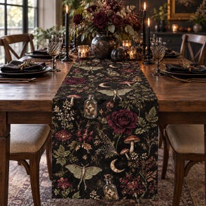 Puede incluir: Un camino de mesa negro con un estampado botánico y celestial que incluye rosas de color rojo oscuro, polillas verdes, setas, telarañas, lunas crecientes y frascos de boticario de cristal llenos de plantas sobre una mesa de madera.