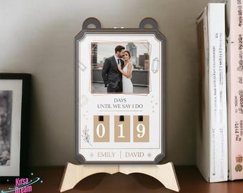 May include: A wooden wedding countdown calendar with a photo of a couple. The calendar displays "DAYS UNTIL WE SAY I DO" and shows "019" days remaining. The names "EMILY" and "DAVID" are printed below the numbers. The calendar is on a wooden stand.