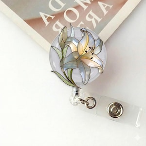 May include: A floral badge reel featuring a lily design with white, yellow, and light blue petals. The badge reel has a clear plastic retractable cord and a white plastic clip. The lily design is on a clear, round background.