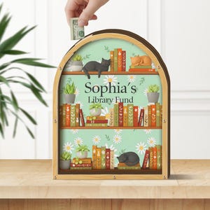May include: Wooden money bank shaped like a bookshelf, with the text "Sophia's Library Fund." The bank features a mint green background with daisies, shelves with books, potted plants, and sleeping cat illustrations. A hand is inserting a dollar bill.