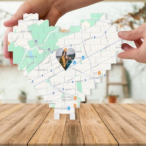 Puede incluir: Una pieza de arte en forma de corazón con un mapa y una foto en el centro. El mapa muestra calles y puntos de referencia, con una foto de dos personas en un recorte en forma de corazón. La pieza está sostenida por manos y descansa sobre una superficie de madera.