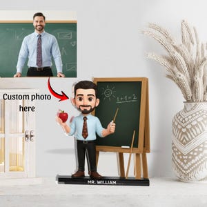 Puede incluir: Figura de profesor personalizada con una pizarra en miniatura y una manzana. La figura es de un hombre con camisa y corbata azul claro, sosteniendo una manzana y un puntero. La pizarra dice "1+1=2". La base dice "MR. WILLIAM".