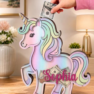 Pu&ograve; includere: Un salvadanaio a forma di unicorno dai colori pastello, con criniera e coda arcobaleno. Il nome "Sophia" &egrave; visualizzato sul davanti e il salvadanaio &egrave; pieno di soldi. Una mano inserisce una banconota da 100 euro nella fessura superiore.