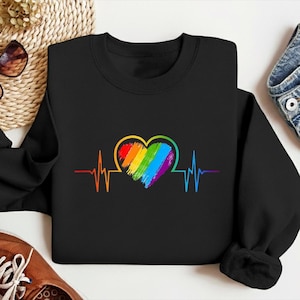 May include: Black sweatshirt featuring a rainbow heart design with a heartbeat line. The heart is filled with vibrant colors, including red, orange, yellow, green, blue, and purple. The design is centered on the front of the sweatshirt.
