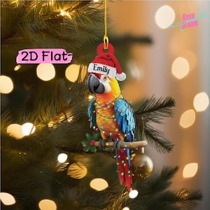 Pode incluir: Um ornamento suspenso plano 2D com um papagaio colorido de penas amarelas, laranja e azuis. O papagaio usa um gorro de Pai Natal vermelho e branco com o nome Emily impresso e está enrolado num fio de luzes de Natal multicoloridas.