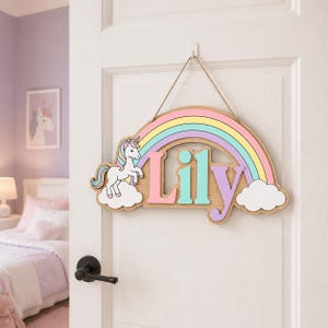 May include: A wooden wall sign featuring a pastel rainbow arch, a white unicorn with a multicoloured mane, and the name Lily in large, raised letters. The sign is suspended by a thin jute rope and includes white cloud accents on both ends.