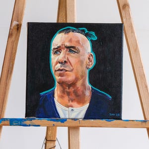 Può includere: Un dipinto di ritratto su tela nera, raffigurante un uomo con occhi blu, piercing e una testa rasata con un piccolo chignon. Il dipinto è delineato in verde acqua e firmato "BMW 25". Il soggetto indossa una camicia blu scuro sopra una maglietta bianca.