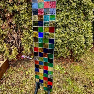 May include: A rectangular stained glass panel with a grid of colorful squares in shades of red, blue, green, and yellow. The panel is supported by a black metal stand and is displayed outdoors in a grassy area.