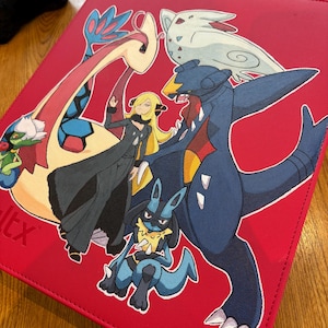 May include: Red binder featuring various Pokémon characters, including Garchomp, Lucario, and Cynthia. The binder has a stitched border and a black plush toy is visible in the background.