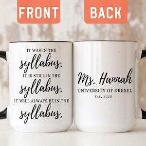 May include: Two white ceramic mugs with black handles and rims. The left mug features the text: IT WAS IN THE syllabus. IT IS STILL IN THE syllabus. IT WILL ALWAYS BE IN THE syllabus. The right mug features: Ms. Hannah UNIVERSITY OF DREXEL Est. 2026.