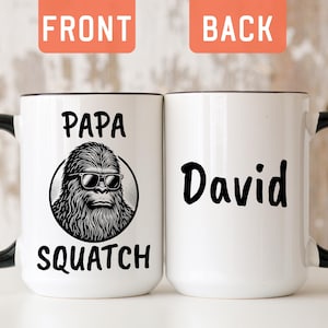 May include: Two white ceramic mugs with black handles and rims. The left mug features a black graphic of a hairy creature wearing sunglasses inside a circle, with the text Papa Squatch. The right mug displays the name David in black lettering.
