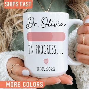 May include: A white ceramic mug featuring the black text Dr. Olivia, a pink horizontal progress bar, the black text IN PROGRESS..., a small pink heart, and the black text EST. 2026. The mug has a curved handle and a smooth, glossy finish.