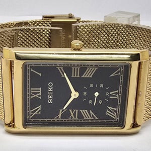 Puede incluir: Un reloj Seiko dorado con una esfera negra rectangular y números romanos dorados. El reloj tiene una correa de malla y una pequeña esfera de segundos. El reloj es de diseño clásico.