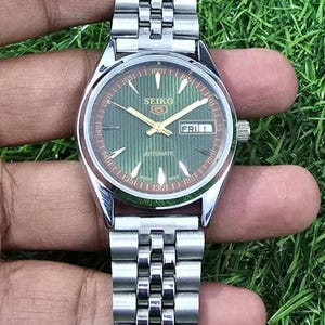 Puede incluir: Un reloj de pulsera autom&aacute;tico Seiko plateado con esfera verde y visualizaci&oacute;n de fecha. El reloj tiene una correa plateada y lo sostiene una mano sobre un fondo verde. La esfera muestra el d&iacute;a de la semana como "FRI 1".