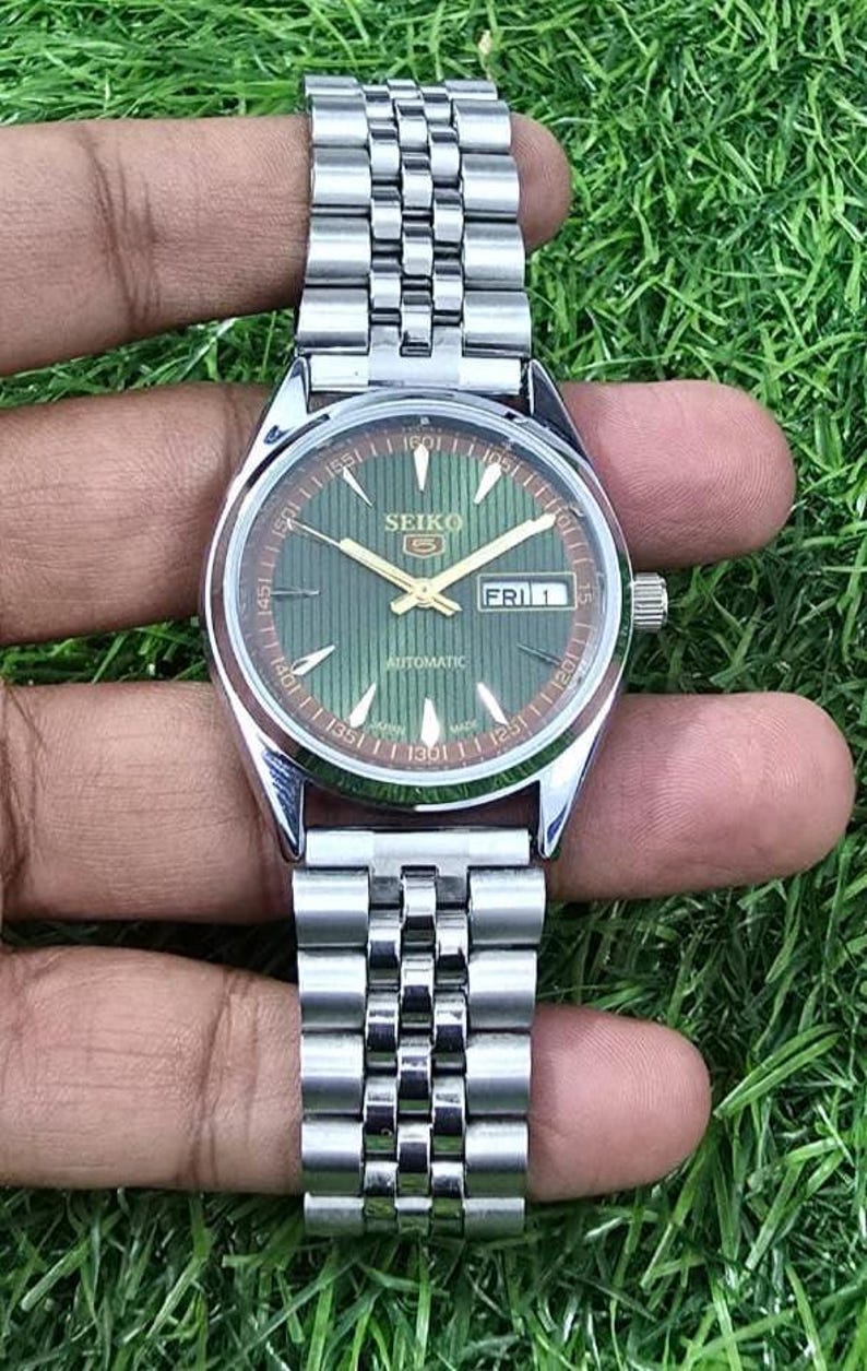 Puede incluir: Un reloj de pulsera autom&aacute;tico Seiko plateado con esfera verde y visualizaci&oacute;n de fecha. El reloj tiene una correa plateada y lo sostiene una mano sobre un fondo verde. La esfera muestra el d&iacute;a de la semana como "FRI 1".