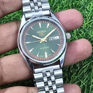 Puede incluir: Un reloj de pulsera autom&aacute;tico Seiko plateado con esfera verde y correa de acero inoxidable. La esfera muestra el d&iacute;a y la fecha, con agujas y marcadores dorados. El reloj se sostiene sobre un fondo verde.