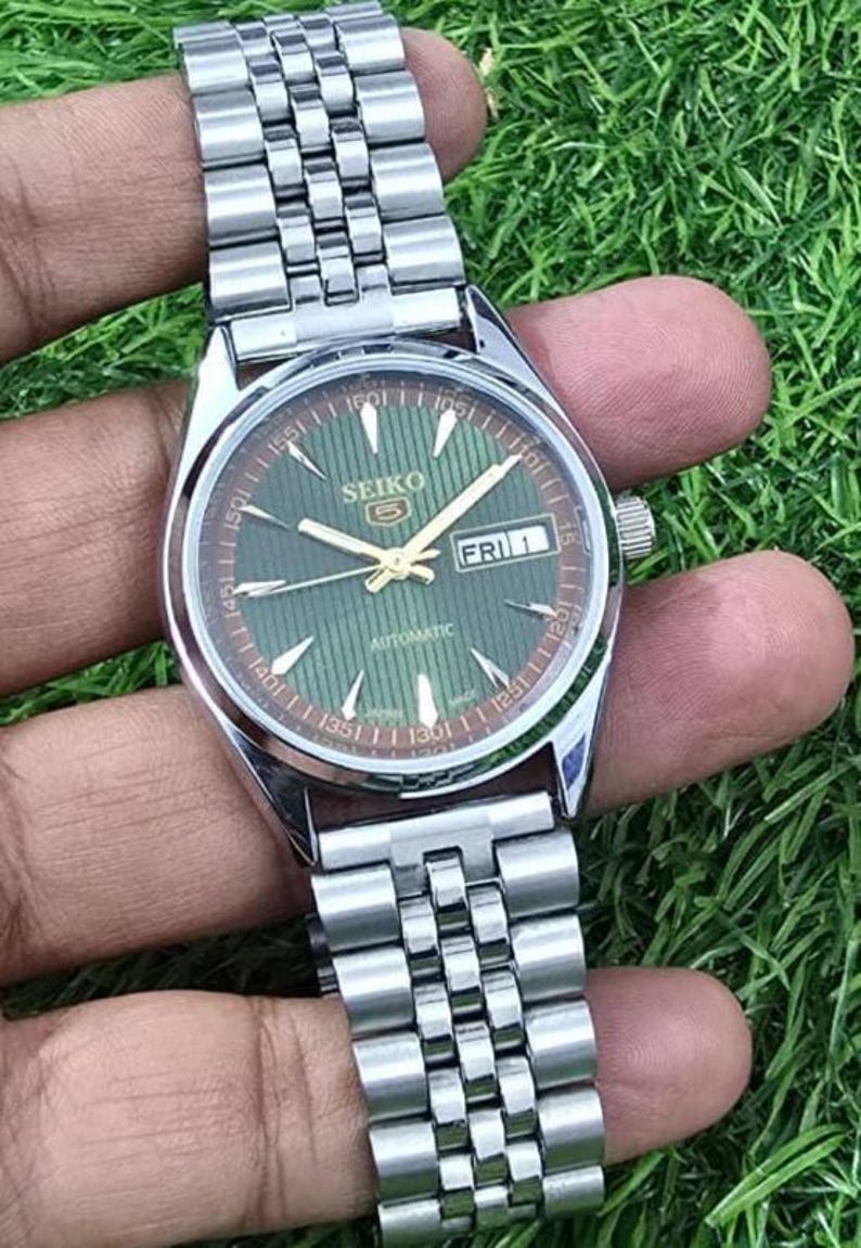 Puede incluir: Un reloj de pulsera autom&aacute;tico Seiko plateado con esfera verde y correa de acero inoxidable. La esfera muestra el d&iacute;a y la fecha, con agujas y marcadores dorados. El reloj se sostiene sobre un fondo verde.