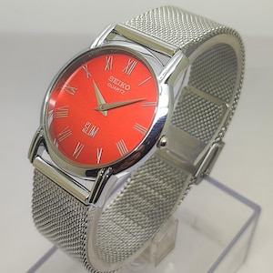 Puede incluir: Un reloj Seiko Quartz plateado con esfera roja brillante y correa de malla plateada. El reloj tiene n&uacute;meros romanos y la palabra "SLIM" en la parte inferior de la esfera. El reloj se exhibe en un soporte transparente.