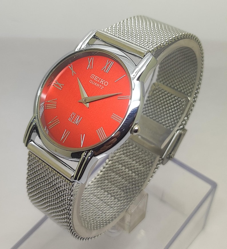 Puede incluir: Un reloj Seiko Quartz plateado con esfera roja brillante y correa de malla plateada. El reloj tiene n&uacute;meros romanos y la palabra "SLIM" en la parte inferior de la esfera. El reloj se exhibe en un soporte transparente.
