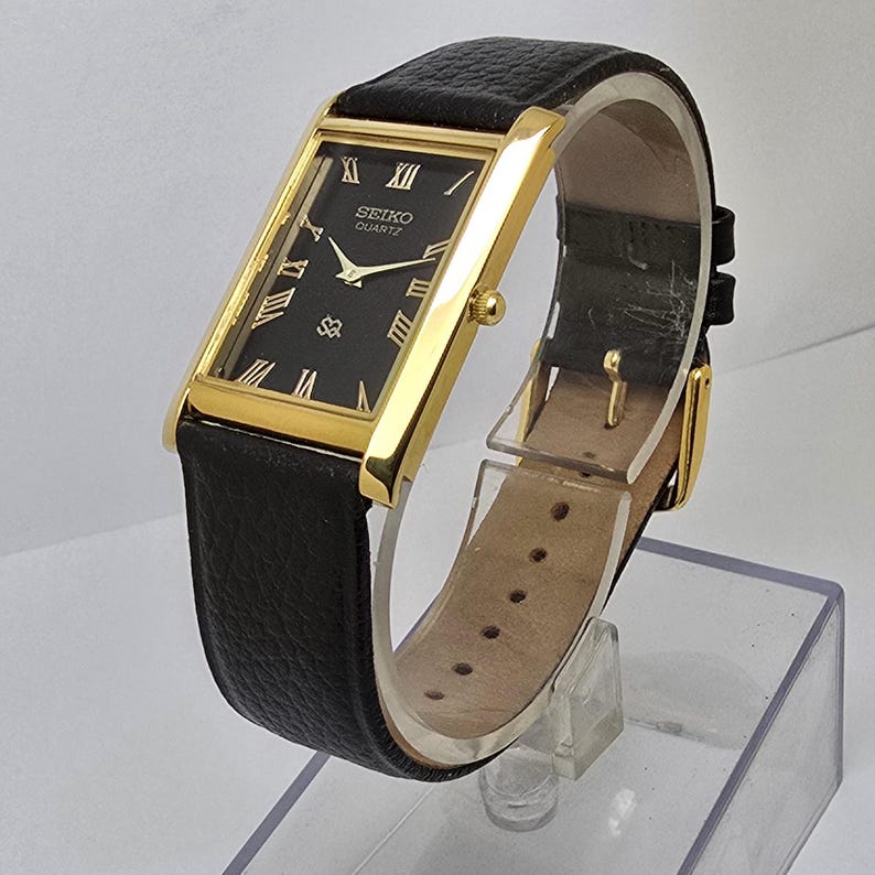 May include: A rectangular gold-toned Seiko Quartz watch with a black face and Roman numeral hour markers. The watch has a black leather band. The watch face displays the Seiko Quartz logo and a small emblem. The watch is displayed on a clear stand.