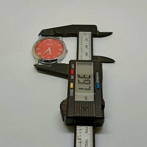 Puede incluir: Un reloj Seiko plateado con esfera roja y n&uacute;meros romanos se mide con un calibrador digital negro. El calibrador muestra una medida de 3,91 mm.