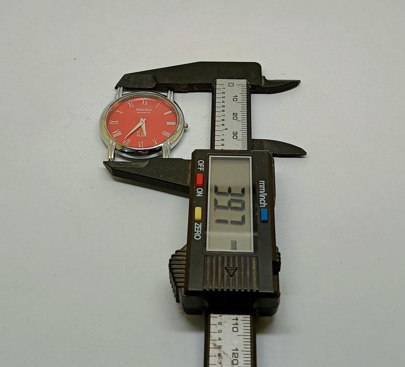 Puede incluir: Un reloj Seiko plateado con esfera roja y n&uacute;meros romanos se mide con un calibrador digital negro. El calibrador muestra una medida de 3,91 mm.
