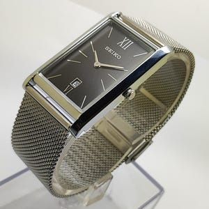 Puede incluir: Un reloj Seiko rectangular con esfera gris y agujas plateadas. El reloj tiene una correa de malla plateada y una pantalla de fecha a las 6 en punto. La esfera del reloj muestra el número XII en la parte superior. El reloj mide unos 3,8 cm de ancho.