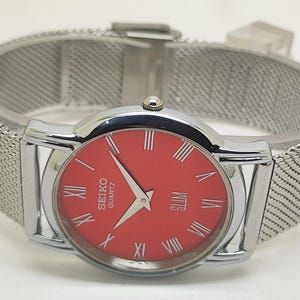 Puede incluir: Un reloj Seiko Quartz plateado con esfera roja y n&uacute;meros romanos. El reloj tiene una correa de malla y un dise&ntilde;o delgado. La esfera del reloj muestra la palabra "SLIM".