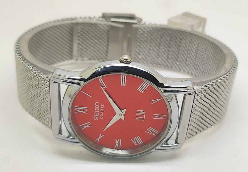 Puede incluir: Un reloj Seiko Quartz plateado con esfera roja y n&uacute;meros romanos. El reloj tiene una correa de malla y un dise&ntilde;o delgado. La esfera del reloj muestra la palabra "SLIM".