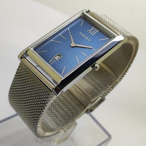 May include: A rectangular Seiko watch with a blue face and silver hands. The watch has a silver mesh strap and a date display at the 6 o'clock position. The watch face has the word "SEIKO" and Roman numeral "XII".