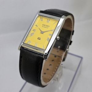Könnte beinhalten: Eine rechteckige Seiko Quartz-Uhr mit goldfarbenem Zifferblatt und römischen Ziffern. Die Uhr hat ein silberfarbenes Gehäuse und ein schwarzes Lederarmband. Das Zifferblatt zeigt den Text "SEIKO QUARTZ" und ein kleines Logo.