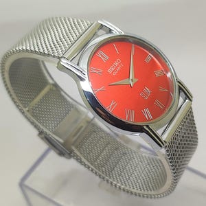 Puede incluir: Reloj Seiko Quartz plateado con esfera roja vibrante y n&uacute;meros romanos. El reloj tiene una correa de malla y un perfil delgado. Un accesorio elegante.