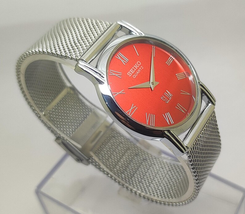 Puede incluir: Reloj Seiko Quartz plateado con esfera roja vibrante y n&uacute;meros romanos. El reloj tiene una correa de malla y un perfil delgado. Un accesorio elegante.