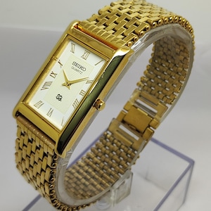 May include: A gold Seiko Quartz watch with a rectangular face and a matching woven metal strap. The watch face is white with black Roman numerals and gold hands. The watch is displayed on a clear stand.