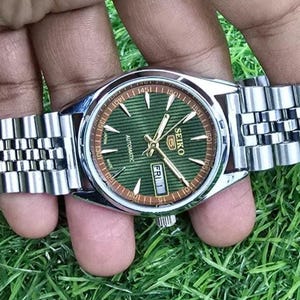 Puede incluir: Un reloj de pulsera autom&aacute;tico Seiko plateado con esfera verde y agujas doradas. El reloj tiene una correa de acero inoxidable y una pantalla de fecha que muestra "VIE 1". El reloj se muestra sobre hierba verde.
