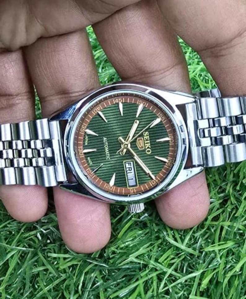Puede incluir: Un reloj de pulsera autom&aacute;tico Seiko plateado con esfera verde y agujas doradas. El reloj tiene una correa de acero inoxidable y una pantalla de fecha que muestra "VIE 1". El reloj se muestra sobre hierba verde.