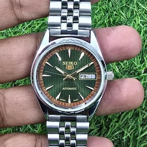 May include: A silver-toned Seiko automatic wristwatch with a green dial and a stainless steel band. The watch face displays the day of the week and the time. The watch is held against a background of green grass.