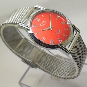 Puede incluir: Un reloj Seiko Quartz plateado con una esfera roja vibrante y n&uacute;meros romanos plateados. El reloj tiene una correa de malla y una caja plateada pulida. La esfera muestra el nombre de la marca y la palabra "Quartz".