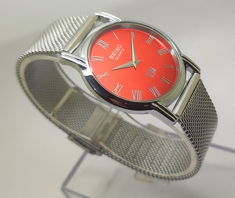 Puede incluir: Un reloj Seiko Quartz plateado con una esfera roja vibrante y n&uacute;meros romanos plateados. El reloj tiene una correa de malla y una caja plateada pulida. La esfera muestra el nombre de la marca y la palabra "Quartz".