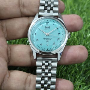 May include: A silver wristwatch with a light blue face and a metal strap. The watch face displays Arabic numerals and the text "hmt Janata PARA SHOCK 17 JEWELS MADE IN INDIA". The watch is shown against a green background.
