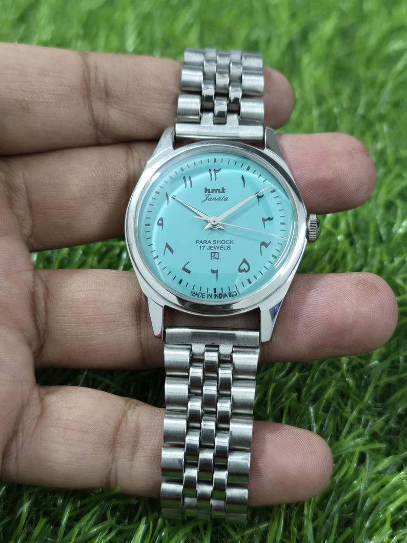 May include: A silver wristwatch with a light blue face and a metal strap. The watch face displays Arabic numerals and the text "hmt Janata PARA SHOCK 17 JEWELS MADE IN INDIA". The watch is shown against a green background.