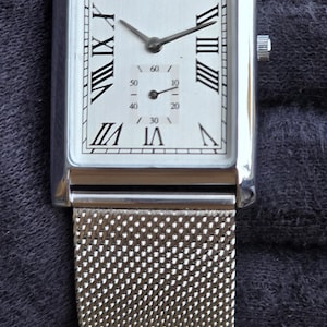 Op de afbeelding: Een rechthoekig Seiko horloge met een zilveren mesh band. De wijzerplaat is wit met zwarte Romeinse cijfers en wijzers. Het horloge heeft een kleine secondenwijzer.
