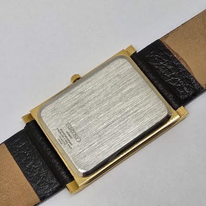 May include: A rectangular gold-toned watch with a silver-colored back and a black leather band. The watch band is attached to the watch face with a gold-toned frame. The watch band is 20 mm wide.