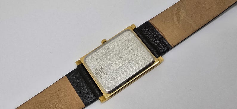 May include: A rectangular gold-toned watch with a silver-colored back and a black leather band. The watch band is attached to the watch face with a gold-toned frame. The watch band is 20 mm wide.