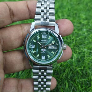 Può includere: Un orologio Citizen Automatic argentato con quadrante verde e cinturino argentato. Il quadrante mostra il giorno e la data, con la scritta "Citizen Automatic" e "21 Jewels". L'orologio è tenuto su uno sfondo verde.