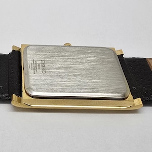May include: A rectangular wristwatch with a gold-toned case and a silver-colored back. The watch is attached to a black and tan leather strap. The watch face is not visible. The watch is a Seiko.