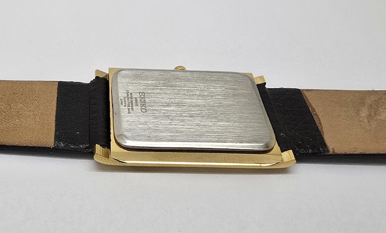 May include: A rectangular wristwatch with a gold-toned case and a silver-colored back. The watch is attached to a black and tan leather strap. The watch face is not visible. The watch is a Seiko.