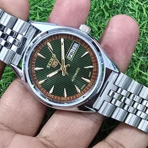 Puede incluir: Un reloj autom&aacute;tico Seiko plateado con esfera verde y agujas doradas. La esfera muestra el d&iacute;a y la fecha, con el d&iacute;a que indica "FRI 1". El reloj tiene una correa plateada y se sostiene sobre un fondo verde.