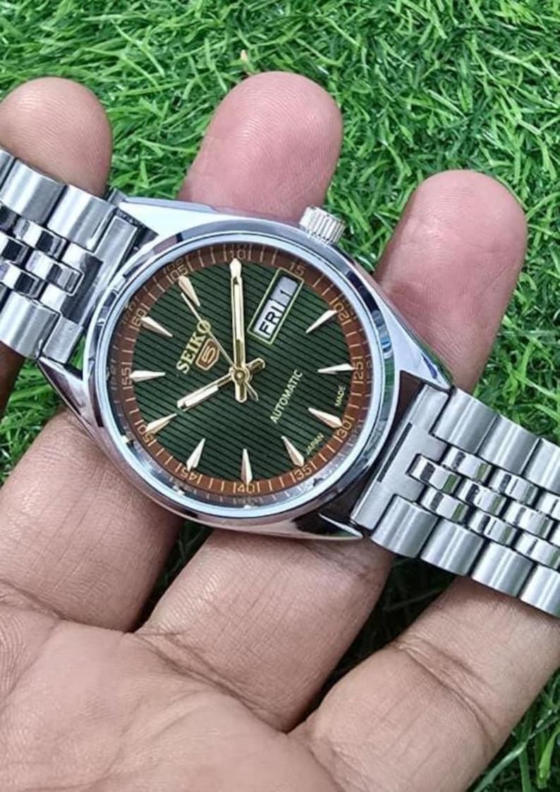 Puede incluir: Un reloj autom&aacute;tico Seiko plateado con esfera verde y agujas doradas. La esfera muestra el d&iacute;a y la fecha, con el d&iacute;a que indica "FRI 1". El reloj tiene una correa plateada y se sostiene sobre un fondo verde.